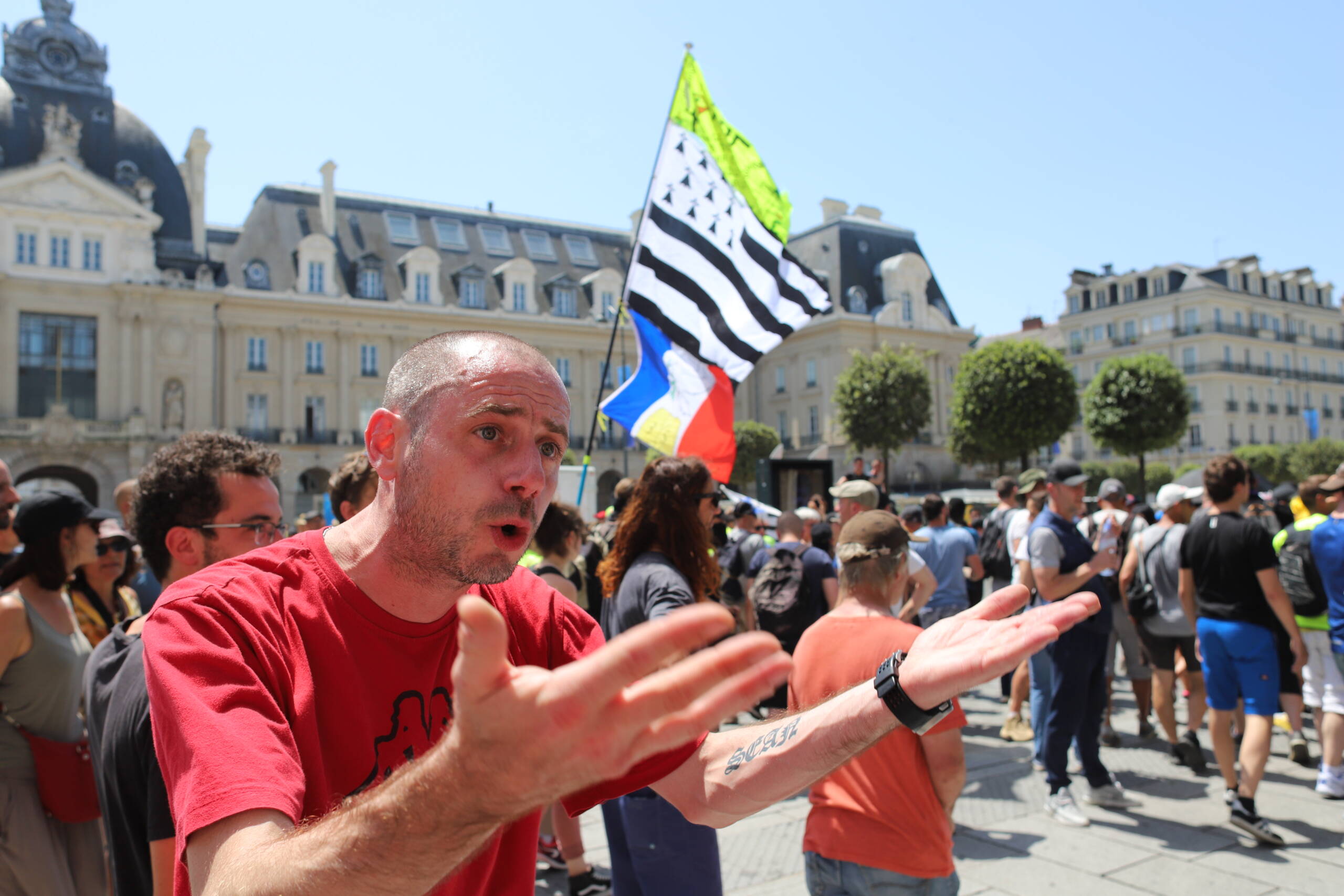 A demonstrator gesturing toward the camera in a sunlit square, with a Breton flag and crowd behind him
