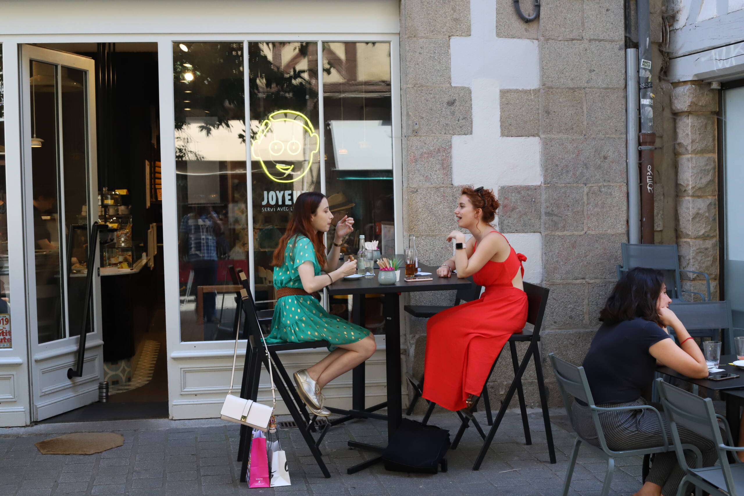 Two women in conversation at a French café table, one in a red dress, one in a teal dress