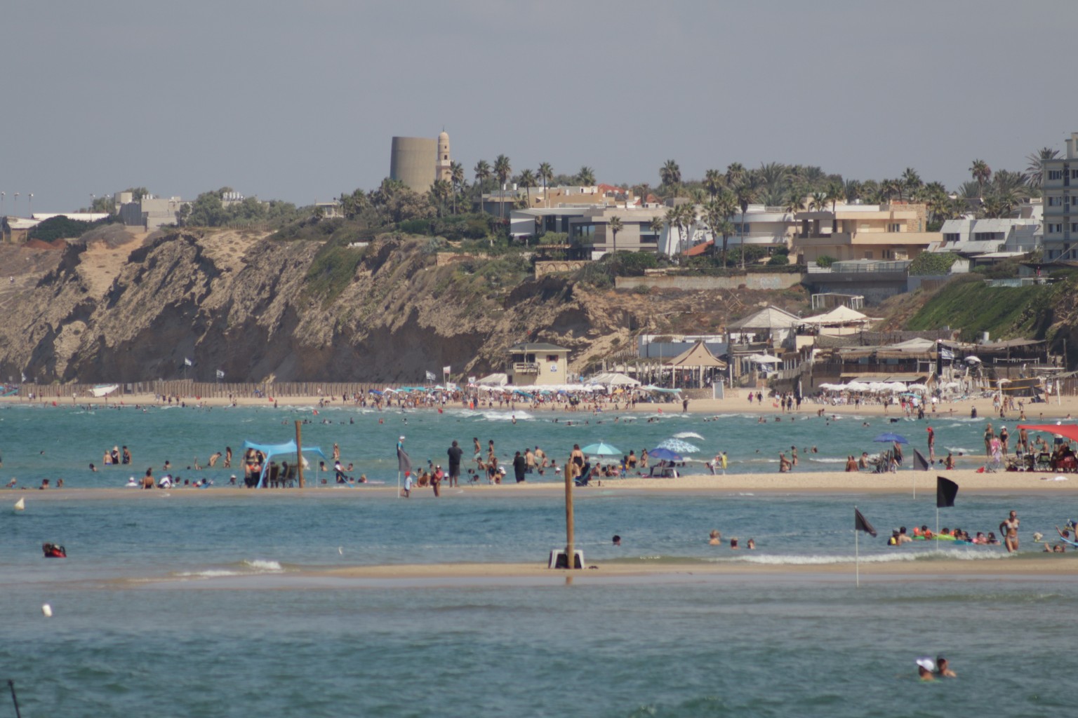 Swimmers and beachgoers along the Herzliya coast, with turquoise water, sandy shallows, parasols and sandstone cliffs beneath palm-lined homes.